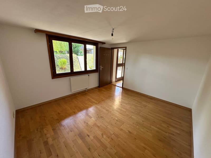 Empty room with wooden flooring, white walls, large window with wooden frame, radiator, and a door leading to another space.