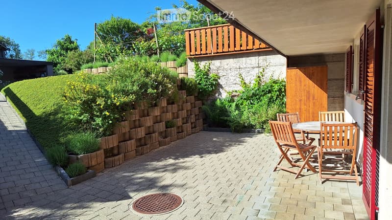 Balcony with a round wooden table and four wooden chairs, surrounded by a brick-paved area, greenery, and a wooden door leading to the house.