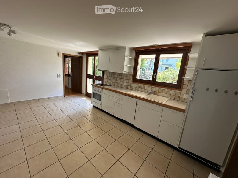 Empty kitchen with tiled floor, white cabinets, a sink, and a refrigerator. Natural light from a large window illuminates the space.