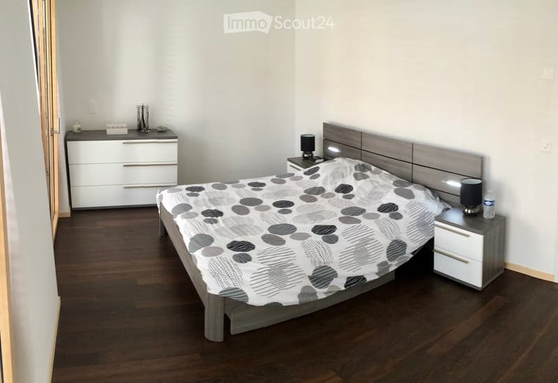 Modern bedroom with a wooden bed, patterned duvet, nightstands, lamps, and a drawer unit.