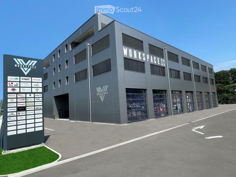 Modern industrial-style building with large glass windows and a sign reading "WORKSPACE II" on the facade. The building has a sleek, contemporary design with a gray exterior and multiple levels.