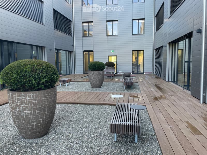 Courtyard with wooden decking, gravel area, benches, tables, and large planters with shrubs.
