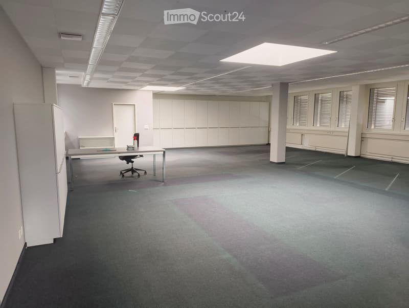 Office space with a table, chair, and white cabinetry. The room has a grey carpeted floor and large windows with blinds.