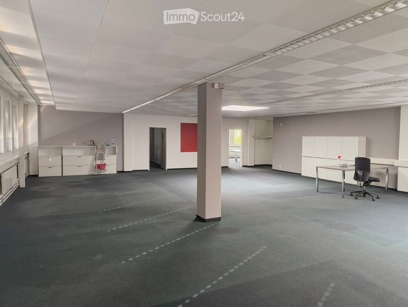 Office space with grey carpet, white walls, ceiling lights, a central pillar, and minimal furniture including a chair, table, and cabinets.