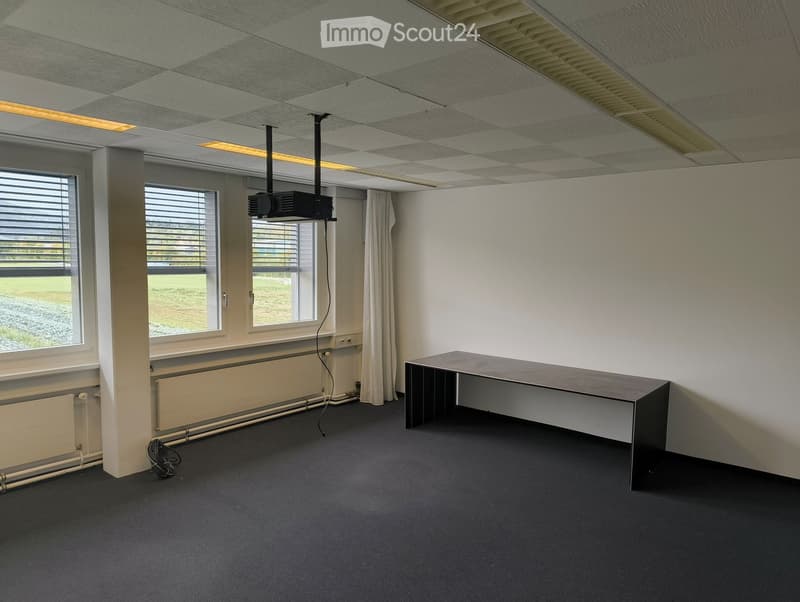 Empty room with a long table, projector, and large windows with blinds. The room has a carpeted floor and white walls.