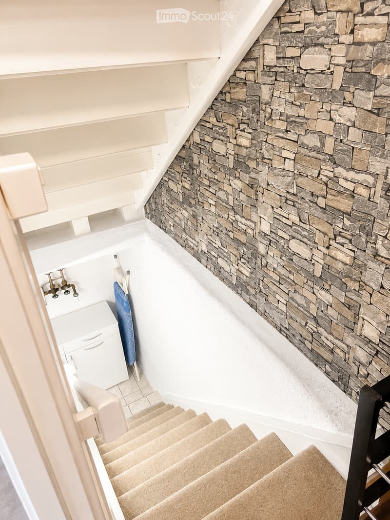 Staircase with beige carpeted steps, white walls, and a stone accent wall on one side. A small white cabinet and a towel are visible in the background.