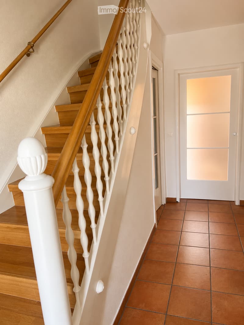 Spacious hallway with wooden staircase featuring white balusters and a wooden handrail, leading to a room with a frosted glass door.