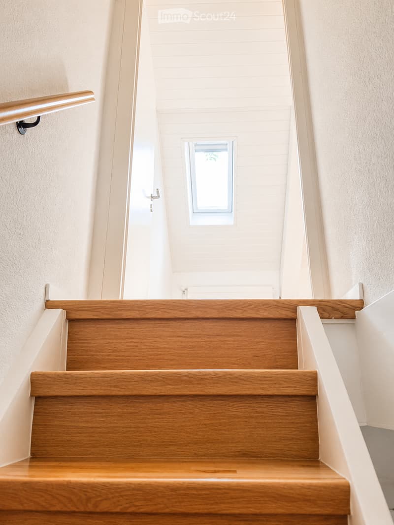 Wooden staircase with a handrail, light-colored walls, and a small window at the top of the stairs.