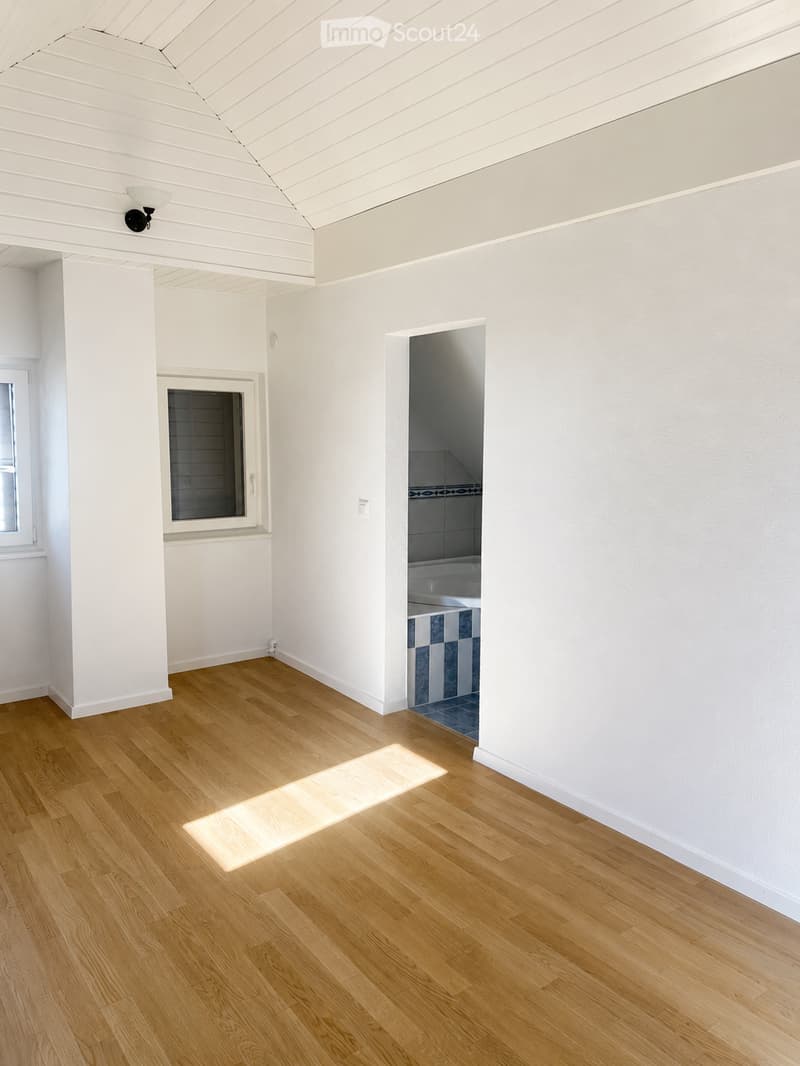 Bright, empty room with light wooden flooring, white walls, and a visible doorway leading to a tiled bathroom area.