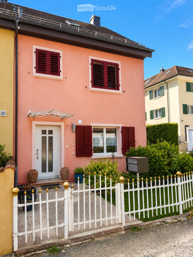 Pink single-story house with white door, red shutters, and decorative vases near the entrance. White picket fence with gold finials surrounds the property.