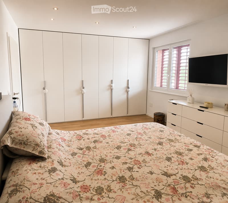 Modern bedroom with a large bed, floral bedspread, white wardrobe, nightstand, television, and decorative items.