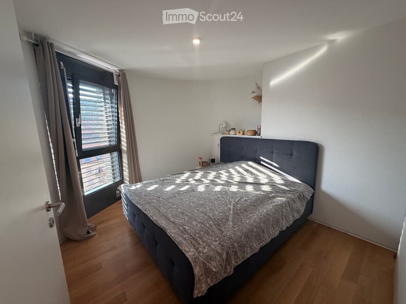 Modern bedroom with wooden flooring, large window with blinds, grey upholstered bed, nightstand with lamp, and decorative items on top.
