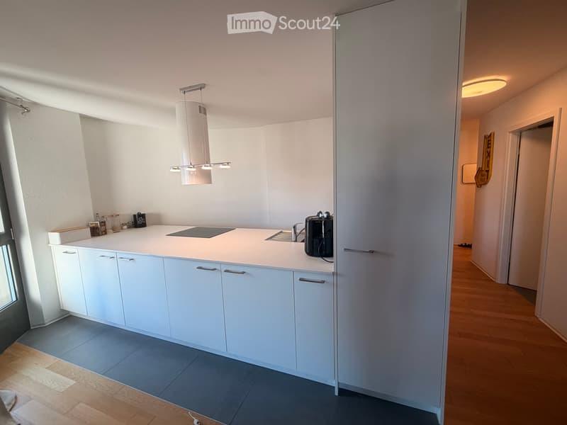 Modern kitchen with white cabinets, light-colored countertop, induction cooktop, sink, and coffee maker. Wooden flooring and pendant light fixture.