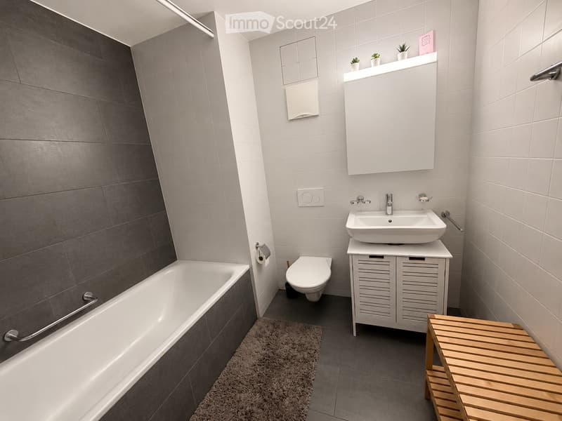 Modern bathroom with a white bathtub, toilet, sink, and a white cabinet with a mirror above it. The walls are tiled in gray and white, and there is a small wooden bench in the corner.