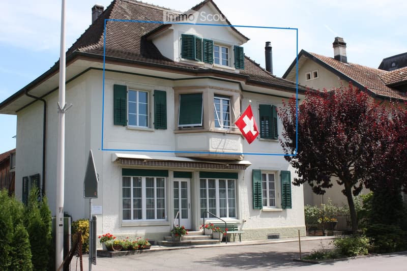 Two-story house with white walls, green shutters, and a red Swiss flag on the balcony. The house features a covered entrance with steps, potted plants, and a bench. The roof is tiled, and there are trees and shrubs in the surrounding garden area.