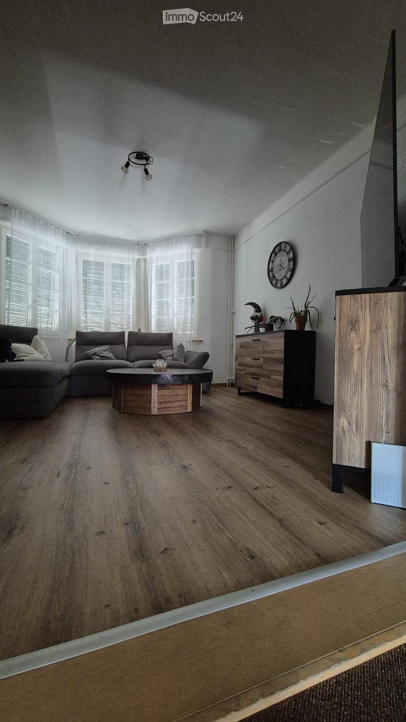 Modern living room with wooden flooring, large windows with white blinds, grey sectional sofa, round wooden coffee table, chest of drawers, wall clock, and decorative plant.