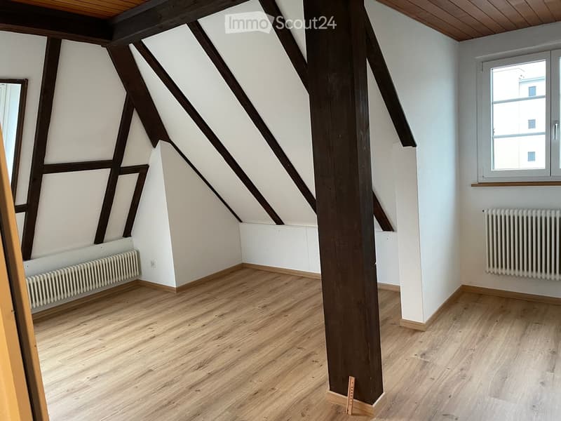 An empty attic room with wooden floors, white walls, exposed wooden beams, and a single window. Radiators are present on two walls.