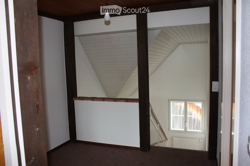 Empty hallway with wooden ceiling beams, white walls, carpeted floor, and a staircase leading upwards. A small window is visible on the right side.