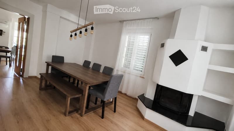 Dining area with wooden table, bench seating, and grey chairs, featuring a modern fireplace and large windows with white shutters.