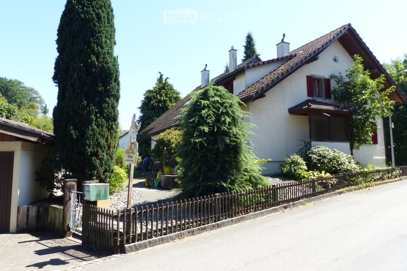 Two-story house with a tiled roof, white exterior, red shutters, and a small garden area with a wooden fence.