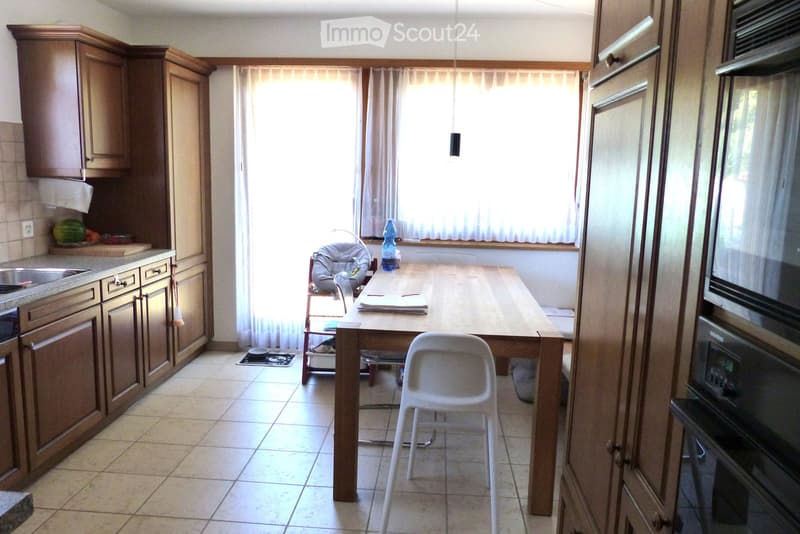The kitchen features wooden cabinets, a central wooden dining table with a white plastic chair, a baby chair, and a bottle on the windowsill. The kitchen includes a sink, an oven, and a refrigerator, with natural light coming through the window with curta