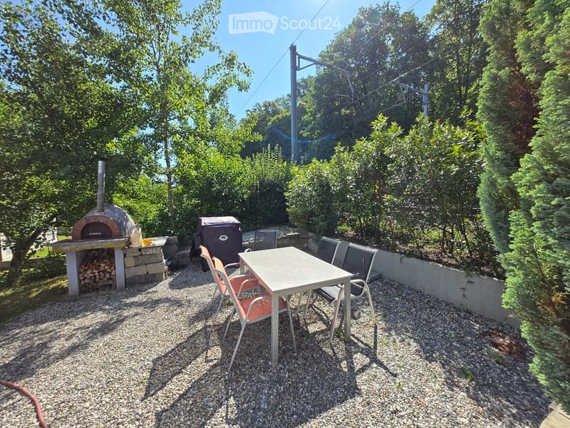 Outdoor patio with a stone pizza oven, a table with chairs, and a covered grill, surrounded by trees and greenery.