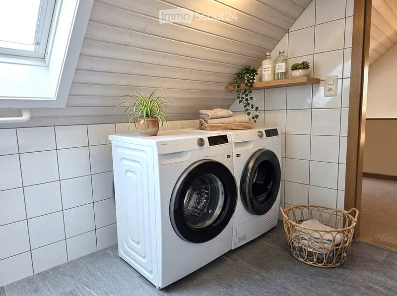 Modern bathroom with white tiled walls, a washing machine and dryer, a wicker basket with towels, and a small potted plant on the counter.
