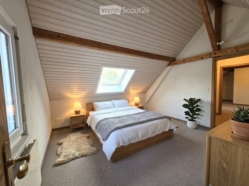 Cozy attic bedroom with wooden slat ceiling, skylight window, bed with white linens, nightstands with table lamps, and potted plants.