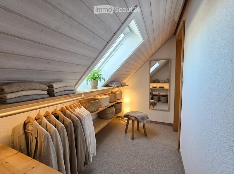 A modern, minimalist walk-in closet with wooden shelves, a clothing rack, woven baskets, a small stool, a mirror, and a potted plant on the windowsill.