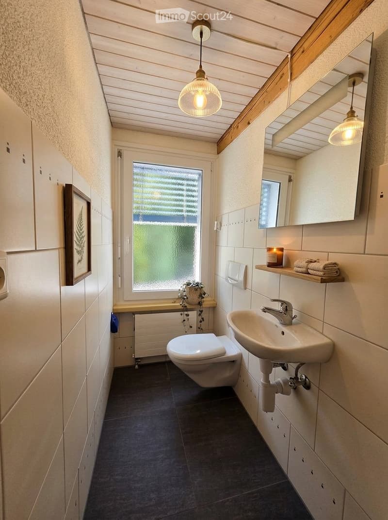 Modern bathroom with a toilet, sink, mirror, and wooden shelf holding towels and a candle. Features a large window with frosted glass and a hanging light fixture.