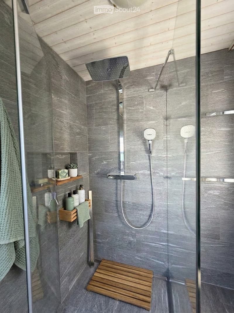 Modern bathroom with grey tiled walls and floor, featuring a large showerhead, handheld shower hose, wooden shower bench, towel, and small bathroom shelf with bottles and a plant.