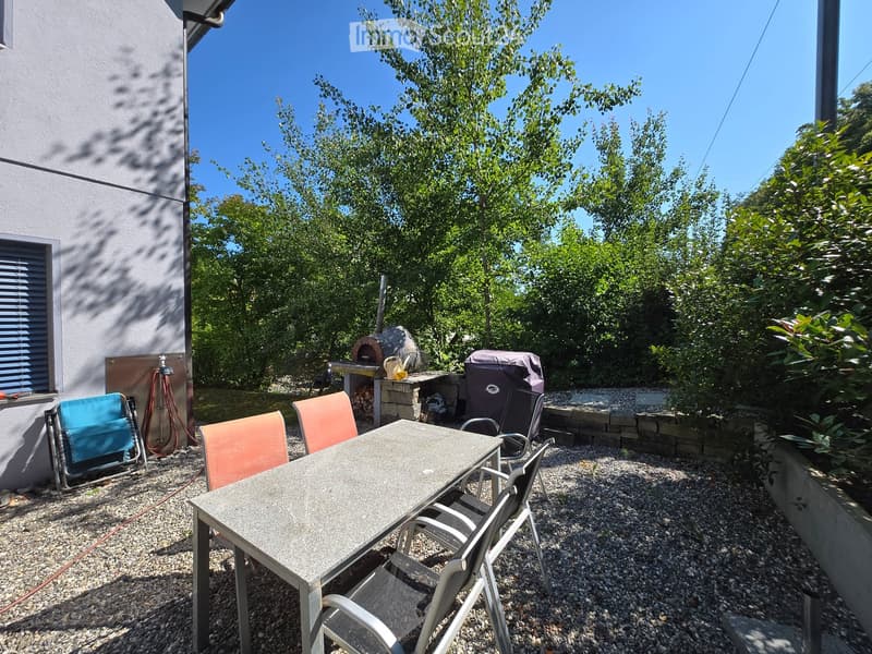 Outdoor patio area with a stone table, four chairs, a grill, and a pizza oven, surrounded by greenery and a stone wall.