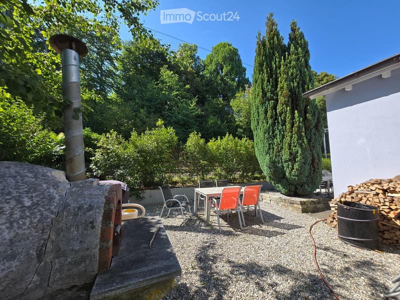 Outdoor patio area with a stone oven, a table with chairs, a barrel, and a stack of firewood, surrounded by trees and greenery.