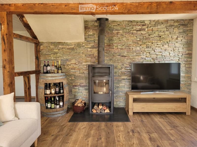 Living room with exposed wooden beams, stone fireplace, wood stove, TV, sofa, and wine barrel holding bottles and glasses.