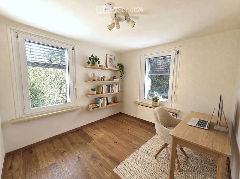 Bright, modern study or home office with wooden flooring, large windows, built-in shelving, desk with laptop and monitor, and a comfortable chair.