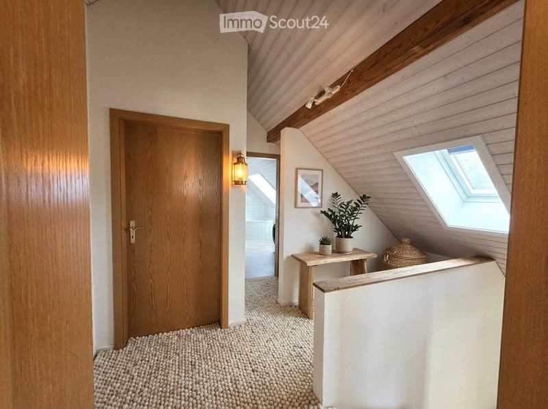 Attic hallway with wooden beams, skylight, small table with potted plants, and a wicker basket. Wooden door leads to a bathroom.