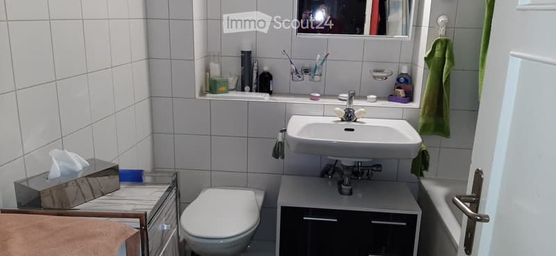 A small bathroom with a white sink, toilet, mirror, and various toiletries on the counter. A green towel hangs on the wall, and a tissue box is placed on a small cabinet.