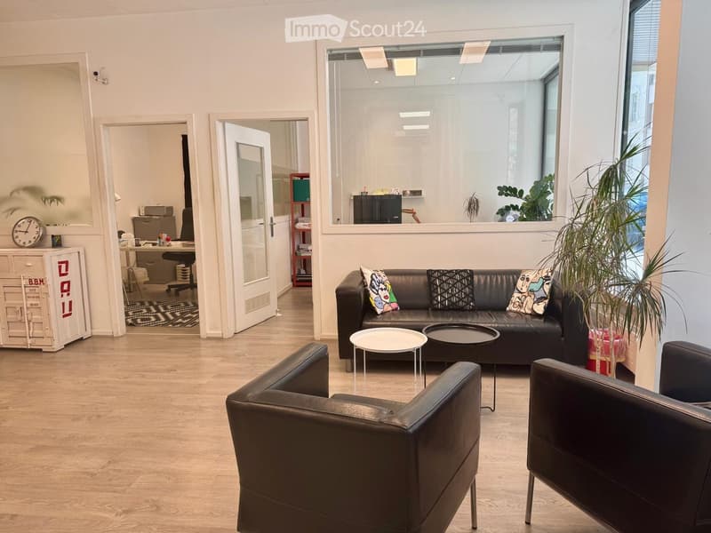 Modern, well-lit room with a black leather sofa, two armchairs, a coffee table, a small round table, a clock, and a screen. The room has large windows and a view of another room with a desk and office equipment.