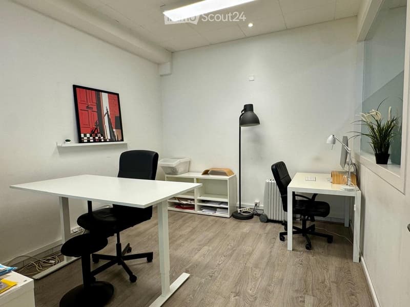 Modern office space with two desks, chairs, computer monitors, keyboards, a floor lamp, a shelf, a radiator, a potted plant, and a picture frame on the wall.