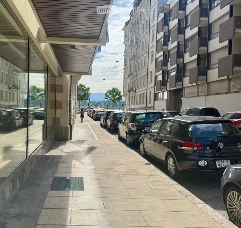 Urban street lined with parked cars, featuring modern buildings with balconies and large windows, a pedestrian walkway, and a clear view of distant mountains.