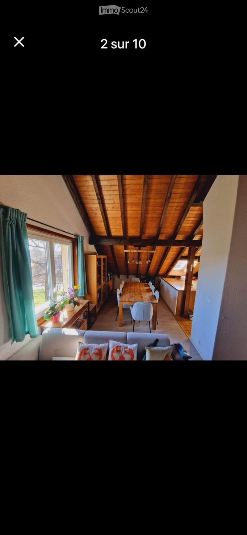 Open-plan living and dining area with exposed wooden beams, large windows, a sofa, coffee table, dining table with chairs, bookshelf, and a small kitchen area.