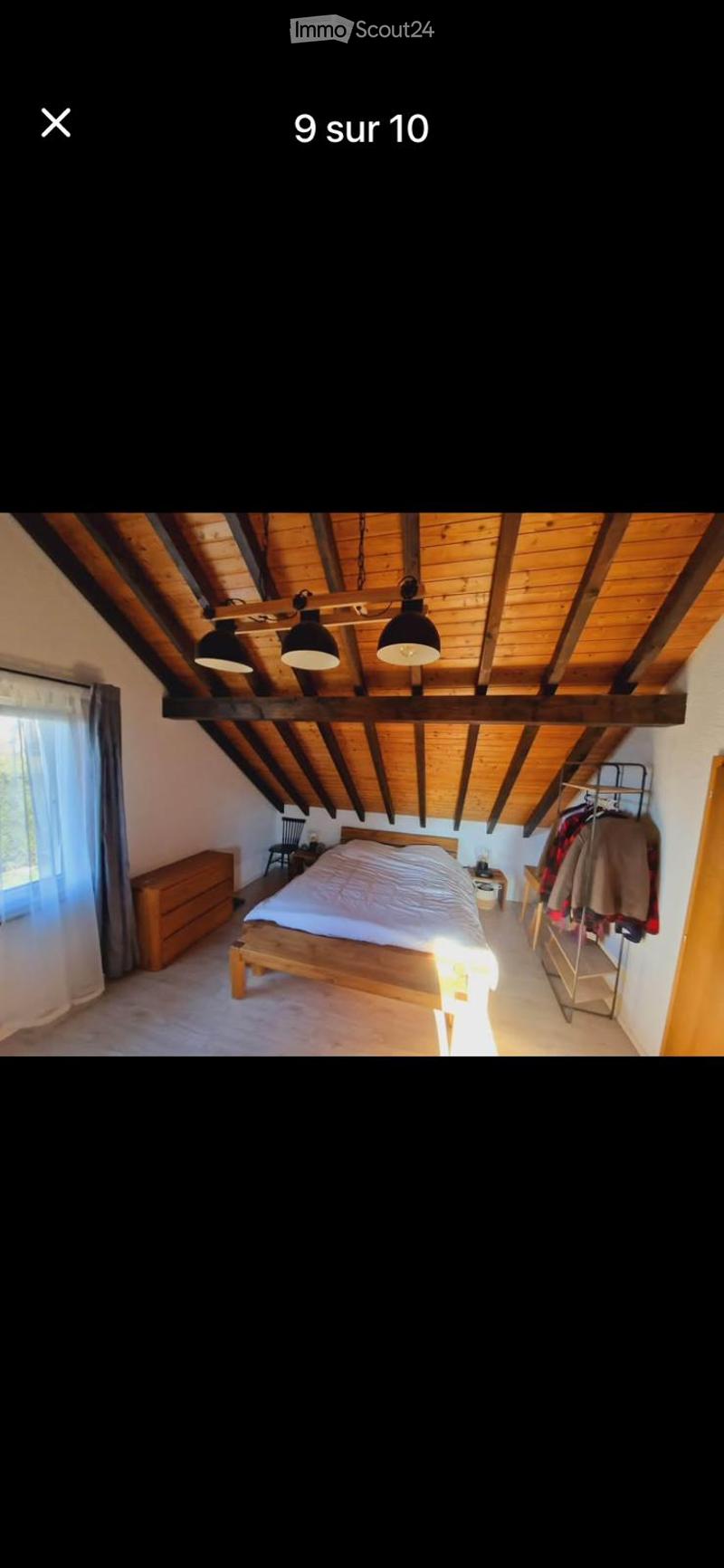 Cozy bedroom with wooden slanted ceiling, exposed beams, and warm lighting. Features a wooden bed, nightstand, chair, and wardrobe with hanging clothes.
