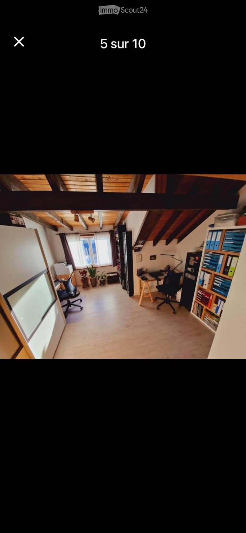 Bright, open room with wooden flooring and slanted wooden ceiling beams. Contains a desk with a chair, a bookcase, a small plant, and a lamp.