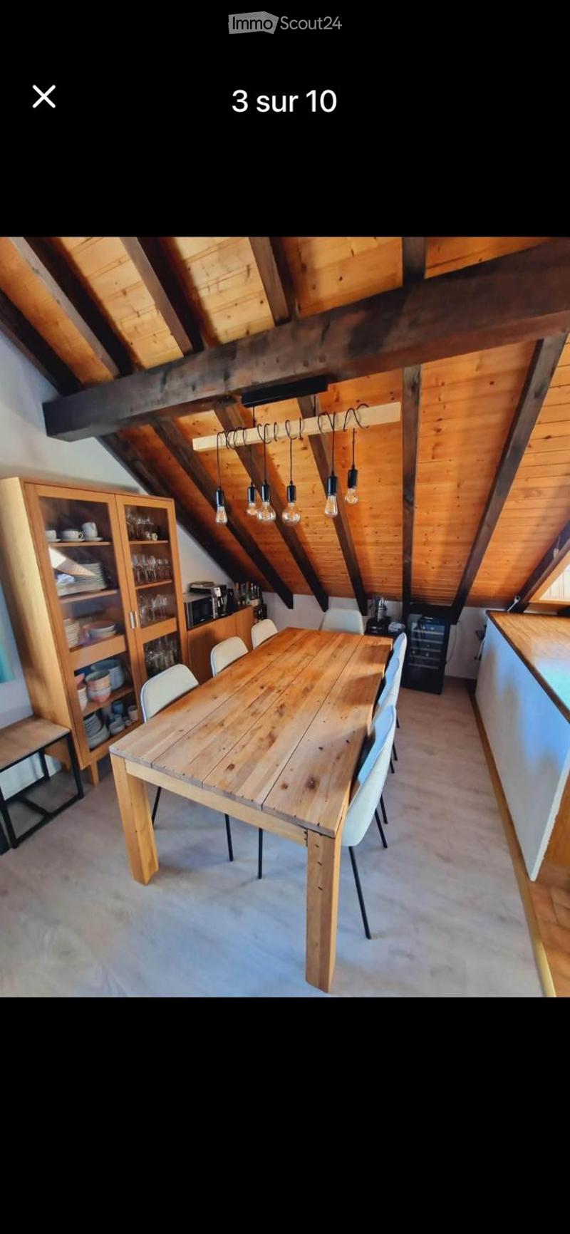 A cozy attic dining area with exposed wooden beams, a large wooden dining table surrounded by white chairs, and built-in wooden cabinets and shelves.