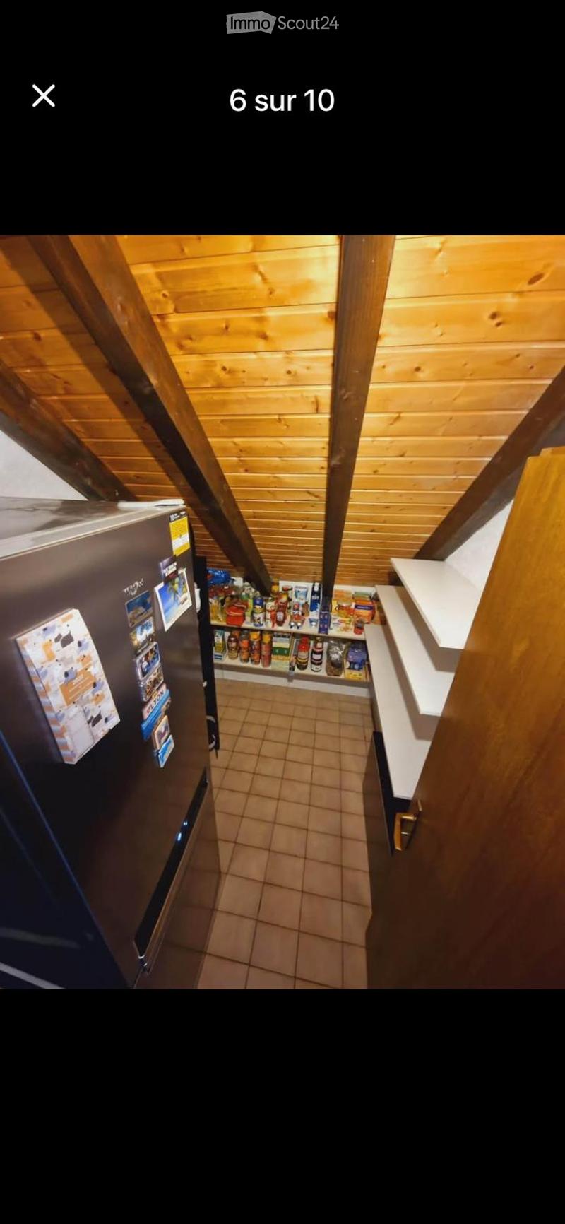 A small kitchen area with wooden walls and ceiling, featuring a refrigerator on the left, shelves with food items, and a tiled floor.