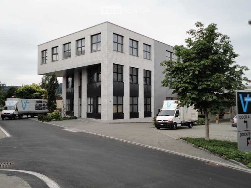 Modern two-story building with white brick facade and large windows, surrounded by a paved area with two delivery trucks and a directional sign.