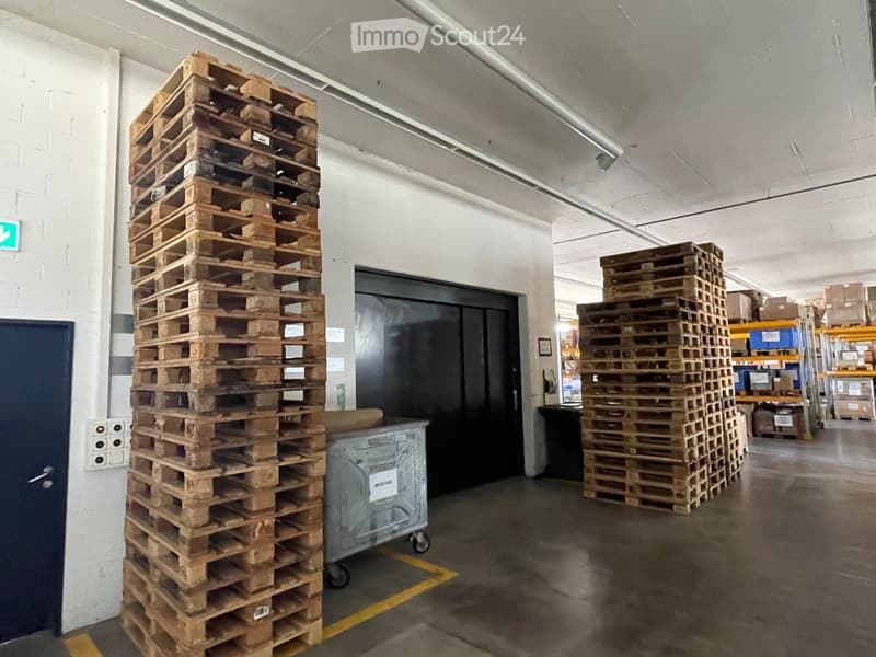 Warehouse space with large stacks of wooden pallets and a metal cart, concrete floor, and industrial lighting.
