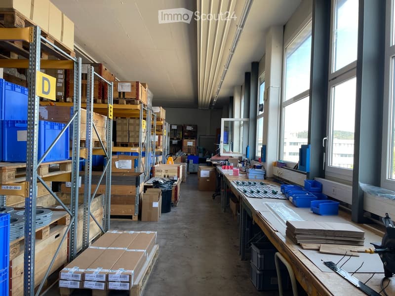 A spacious workshop or storage area with metal shelving units filled with boxes and materials, a workbench with tools and supplies, and large windows allowing natural light.
