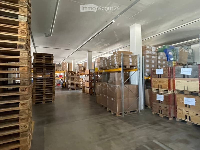 Warehouse space with stacked wooden pallets and cardboard boxes, metal shelving units, and industrial lighting.