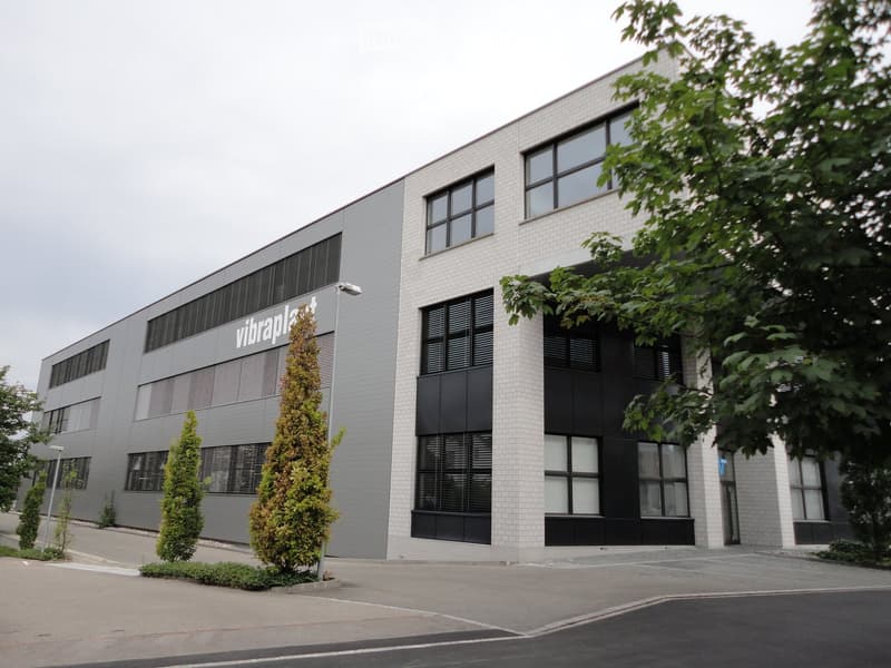 Modern two-story building with white brick facade, large windows, and a sign reading "vibraplast".